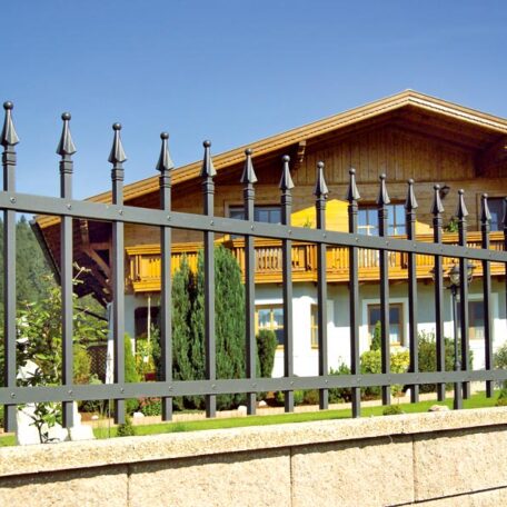 Traditionelles Haus mit Holzbalkon hinter modernem Metallzaun, blauer Himmel, sonniger Tag. HANSEL.kraus metalltechnik gmbh