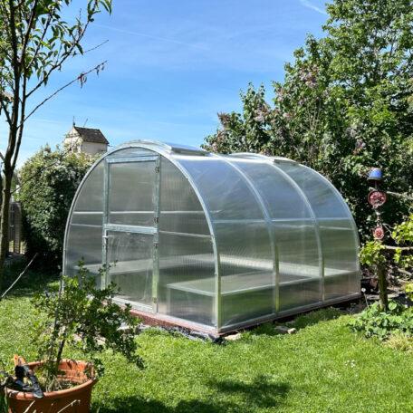 Gewächshaus mit transparenter Runddach-Konstruktion steht im sonnigen Garten unter blauem Himmel. de HANSEL.kraus metalltechnik gmbh