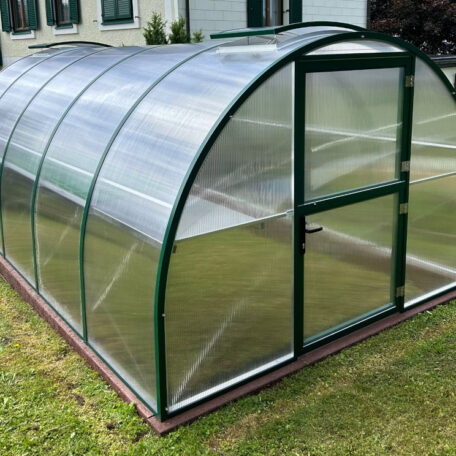 Gewölbtes Gewächshaus aus transparentem Polycarbonat steht auf Rasen vor einem Wohnhaus. HANSEL.kraus metalltechnik gmbh