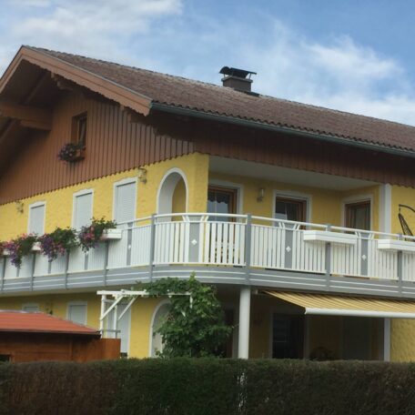 Gelbes Einfamilienhaus mit Balkon, weißen Fensterläden und Blumenkästen unter blauem Himmel. HANSEL.kraus metalltechnik gmbh