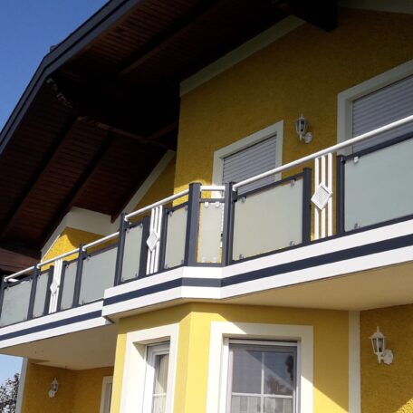 Zweistöckiges gelbes Haus mit modernem Glasbalkon und Holzdach, fotografiert bei Sonnenschein. HANSEL.kraus metalltechnik gmbh