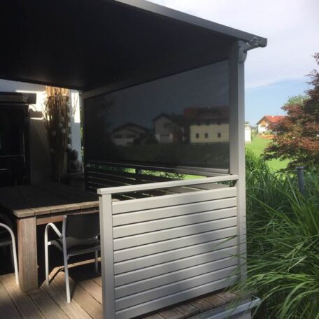 Überdachte Terrasse mit Holzmöbeln und Sichtschutz neben grünem Garten, Wohnhäuser im Hintergrund. HANSEL.kraus metalltechnik gmbh