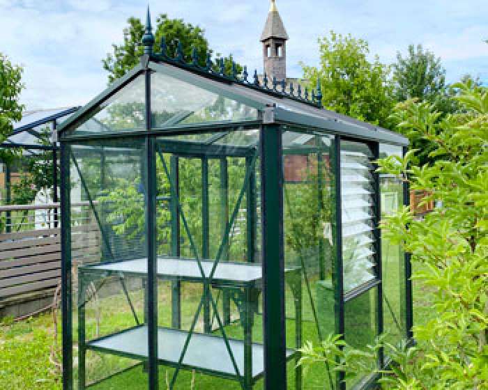 Ein kleines Gewächshaus aus Glas steht im sonnigen Garten, Bäume und ein Kirchturm im Hintergrund. HANSEL.kraus metalltechnik gmbh