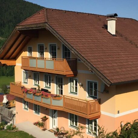 Ein gelbes Haus mit Holzbalkonen und rotem Dach vor bewaldetem Berghang. de HANSEL.kraus metalltechnik gmbh