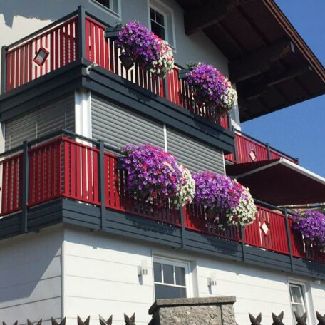 Balkon mit roten Geländern, Blumenkästen voller violetter Blumen an Hausfassade. HANSEL.kraus metalltechnik gmbh