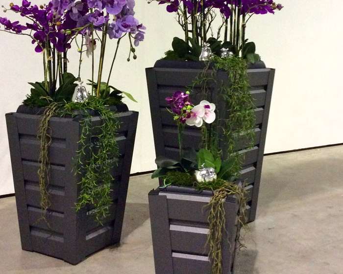 Drei hohe, moderne Blumentöpfe mit lila Orchideen und hängenden Pflanzen auf hellem Boden. de HANSEL.kraus metalltechnik gmbh