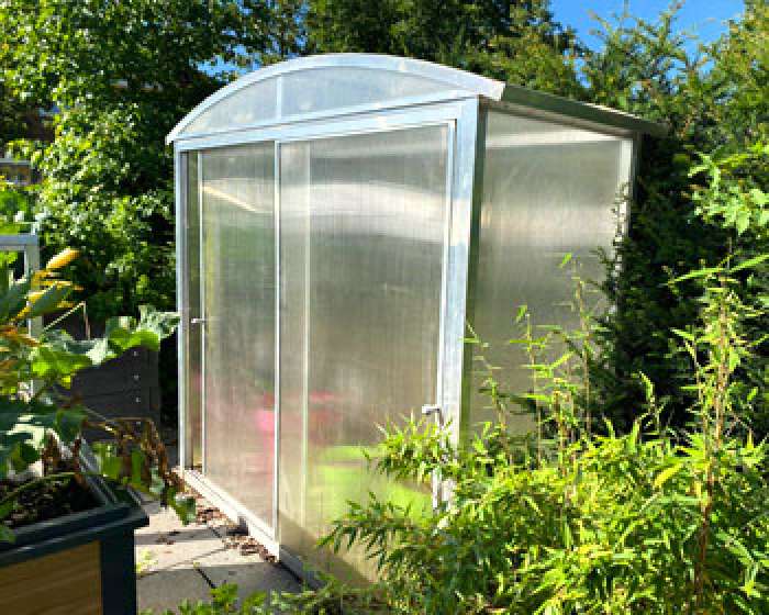 Kleines Gewächshaus mit transparenten Wänden im sonnigen Garten, umgeben von grünen Pflanzen. HANSEL.kraus metalltechnik gmbh