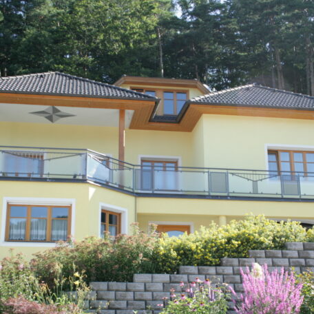 Modernes gelbes Einfamilienhaus mit großem Balkon, Garten und Steinmauer am Waldrand. HANSEL.kraus metalltechnik gmbh