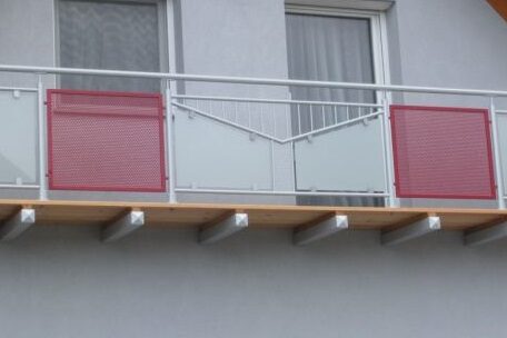 Moderner Balkon mit roten und weißen Sichtschutzelementen vor einem Fenster, Hausfassade sichtbar. HANSEL.kraus metalltechnik gmbh