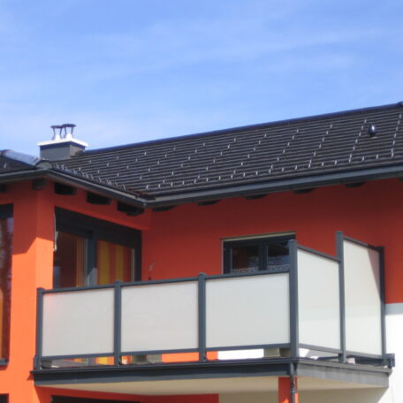 Modernes Wohnhaus mit roter Fassade, Balkon und Solaranlage auf dem Dach bei blauem Himmel. HANSEL.kraus metalltechnik gmbh