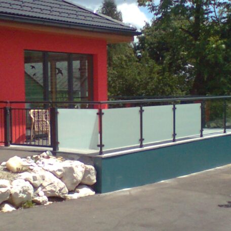 Moderner Balkon mit Glasgeländer an rotem Haus, weiße Steine davor, grüner Garten im Hintergrund. HANSEL.kraus metalltechnik gmbh