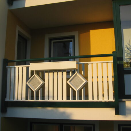 Balkon mit gelber Wand, weißem Geländer und zwei quadratischen Deko-Elementen, sonnige Beleuchtung. HANSEL.kraus metalltechnik gmbh