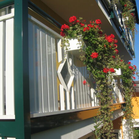 Blumenkasten mit roten Geranien hängt an weißem Balkon einer modernen Hausfassade. de HANSEL.kraus metalltechnik gmbh