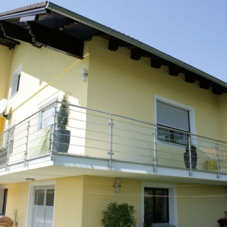 Zweistöckiges gelbes Haus mit modernem Glasbalkon und Pflanzen, blauer Himmel. HANSEL.kraus metalltechnik gmbh