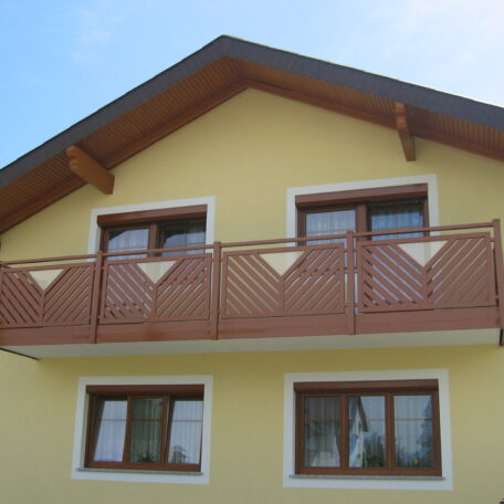 Gelbes Einfamilienhaus mit braunem Holzbalkon und vier Fenstern, blauer Himmel. HANSEL.kraus metalltechnik gmbh