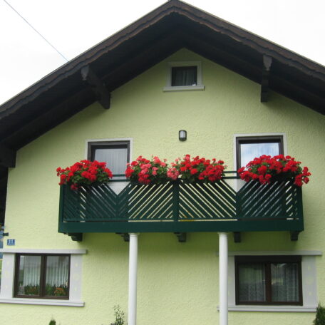 Grünes Einfamilienhaus mit Holzbalkon und roten Blumen, typisch für ländliche Architektur. HANSEL.kraus metalltechnik gmbh