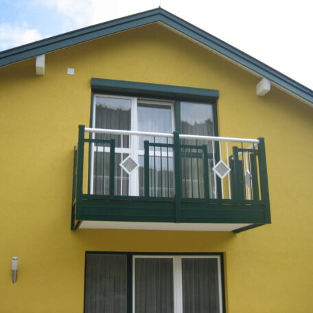 Obergeschoss eines gelben Hauses mit grünem Balkon und weißen Fenstertüren, Tageslicht. HANSEL.kraus metalltechnik gmbh
