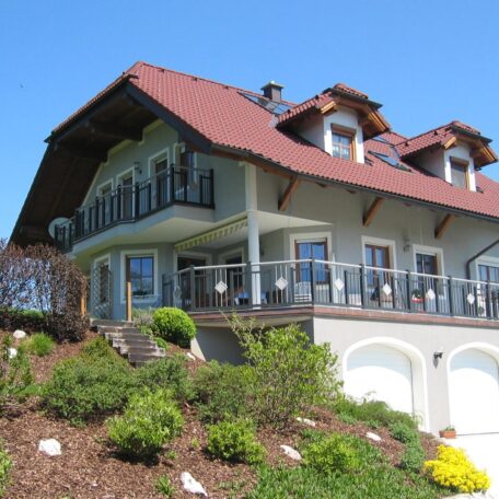 Großes Einfamilienhaus mit rotem Dach, Balkon und gepflegtem Garten an einem sonnigen Hang. HANSEL.kraus metalltechnik gmbh