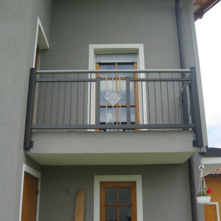 Kleiner Balkon mit Metallgeländer an grauer Hausfassade, darunter Holztür, darüber Fenster. HANSEL.kraus metalltechnik gmbh