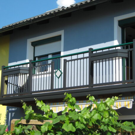 Moderner Balkon mit Metallgeländer an einem blauen Haus, umgeben von grünen Pflanzen. HANSEL.kraus metalltechnik gmbh
