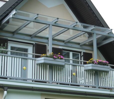 Zweigeschossiger Balkon mit Glasüberdachung und Blumenkästen an Wohnhausfassade, Giebel im Hintergrund. HANSEL.kraus metalltechnik gmbh
