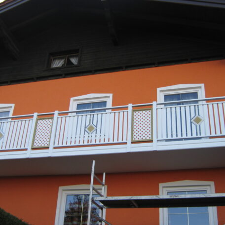 Orange Hausfassade mit weißem Balkon und drei Fenstern, Baugerüst im Vordergrund. de HANSEL.kraus metalltechnik gmbh