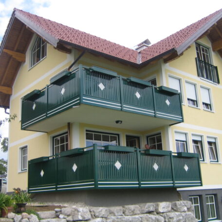 Gelbes Mehrfamilienhaus mit grünem Holzbalkon und Satteldach vor bewölktem Himmel. HANSEL.kraus metalltechnik gmbh