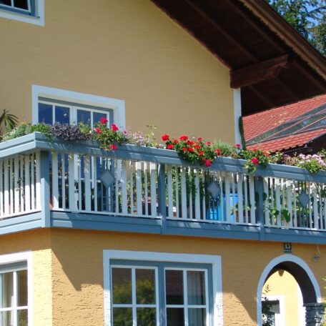 Gelbes Haus mit blauem Balkon, bepflanzt mit bunten Blumen, unter klarem Himmel. de HANSEL.kraus metalltechnik gmbh