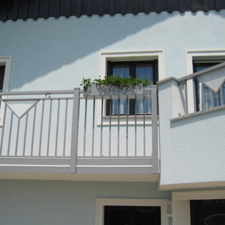 Balkon mit silbernem Geländer und Blumenkasten vor weißer Hausfassade im Sonnenlicht. HANSEL.kraus metalltechnik gmbh