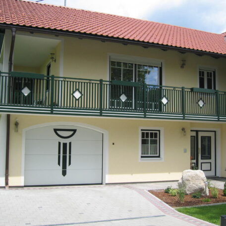 Zweistöckiges Haus mit Balkon, grünem Geländer, Garage und gepflegtem Vorgarten unter rotem Ziegeldach. HANSEL.kraus metalltechnik gmbh