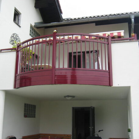 Roter Balkon mit Metallgeländer an weißem Haus, Terrasse darunter, bepflanzt. de HANSEL.kraus metalltechnik gmbh