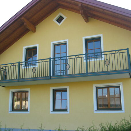 Gelbes Einfamilienhaus mit grünem Balkon, Holzdach und mehreren Fenstern, Frontansicht. HANSEL.kraus metalltechnik gmbh