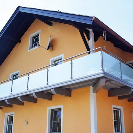 Gelbes Einfamilienhaus mit großzügigem Balkon und moderner Glasbrüstung unter blauem Dach. HANSEL.kraus metalltechnik gmbh