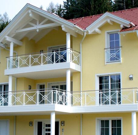 Gelbes Einfamilienhaus mit rotem Dach, mehreren Balkonen und weißen Geländern vor Waldkulisse. HANSEL.kraus metalltechnik gmbh