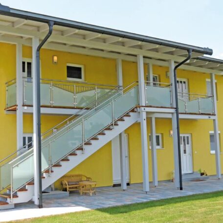 Zweistöckiges gelbes Wohngebäude mit Außentreppe, Balkon und Eingängen, umgeben von Rasenfläche. HANSEL.kraus metalltechnik gmbh