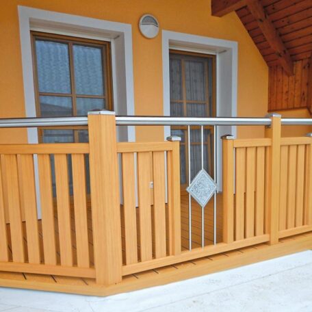 Holzbalkon mit Edelstahlgeländer vor orange gestrichener Hausfassade und zwei Fenstern, Aufnahme bei Tageslicht. HANSEL.kraus metalltechnik gmbh