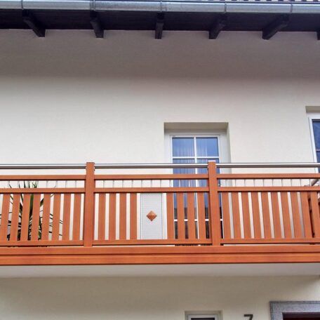 Holzbalkon mit senkrechten Streben vor zwei Fenstern an weißer Hausfassade. de HANSEL.kraus metalltechnik gmbh