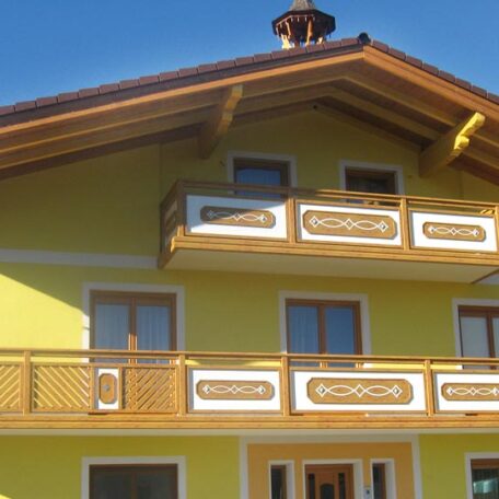 Gelbes Haus mit Holzbalkon und verziertem Giebel unter blauem Himmel. HANSEL.kraus metalltechnik gmbh