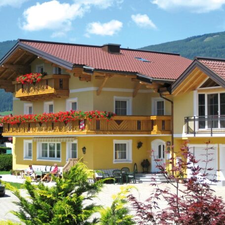 Gelbes Haus mit Holzbalkon und Blumen, umgeben von Bergen und blühenden Sträuchern. HANSEL.kraus metalltechnik gmbh