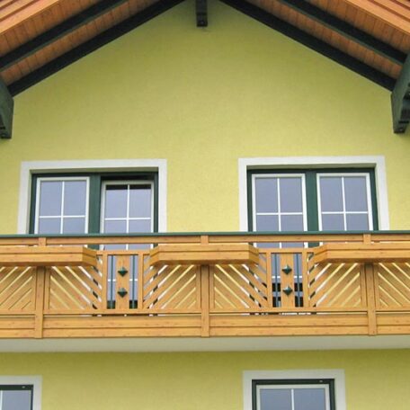 Obergeschoss eines gelben Hauses mit Holzbalkon und zwei Fenstern, blauer Himmel. HANSEL.kraus metalltechnik gmbh