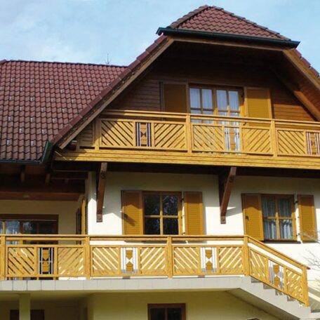Zweistöckiges Haus mit Holzverkleidung, Balkon und geschlossenen Fensterläden unter blauem Himmel. HANSEL.kraus metalltechnik gmbh