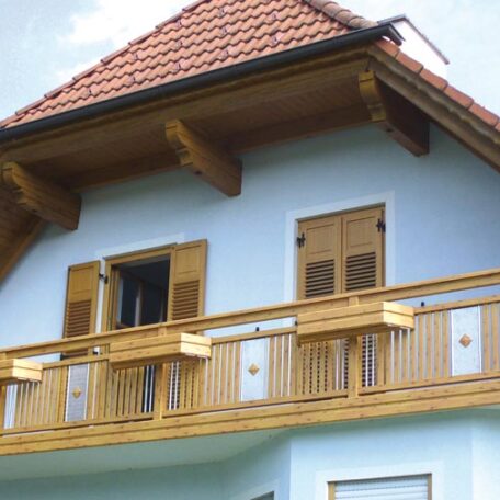 Zweistöckiges Haus mit Balkon, Holzfassade und roten Dachziegeln, blauer Himmel im Hintergrund. HANSEL.kraus metalltechnik gmbh