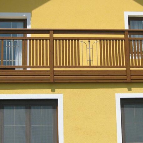 Holzbalkon mit Geländer an gelber Hausfassade, darunter zwei große Fenster, Tageslicht. de HANSEL.kraus metalltechnik gmbh