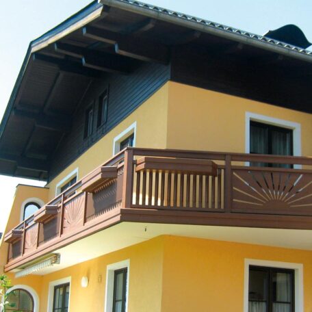 Gelbes Haus mit Holzbalkon und schwarzem Dach vor blauem Himmel, sonniger Tag. HANSEL.kraus metalltechnik gmbh