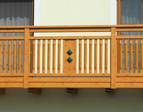 Holzbalkon mit senkrechten Streben und zwei grünen Rauten vor hellgelber Hauswand. HANSEL.kraus metalltechnik gmbh