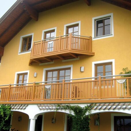 Gelbes Wohnhaus mit Holzbalkonen, zwei Etagen und begrüntem Eingangsbereich, Tageslicht. HANSEL.kraus metalltechnik gmbh