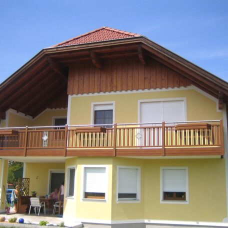 Gelbes Einfamilienhaus mit Holz-Balkon und rotem Satteldach vor blauem Himmel. HANSEL.kraus metalltechnik gmbh