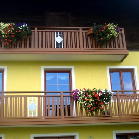 Zwei Holzbalkone mit Blumen an gelber Hausfassade, zentrale Glasfenster und Holztüren. de HANSEL.kraus metalltechnik gmbh