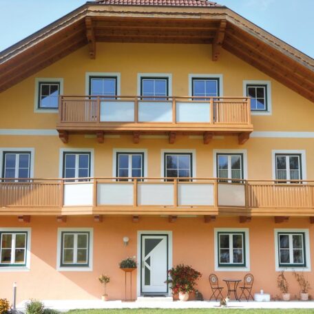 Vierstöckiges gelbes Haus mit Holzbalkonen, Stühlen und Pflanzen vor dem Eingang. de HANSEL.kraus metalltechnik gmbh