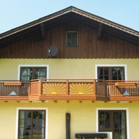 Zweistöckiges gelbes Haus mit Holzfassade, Balkon und weiß gerahmten Fenstern bei sonnigem Wetter. HANSEL.kraus metalltechnik gmbh